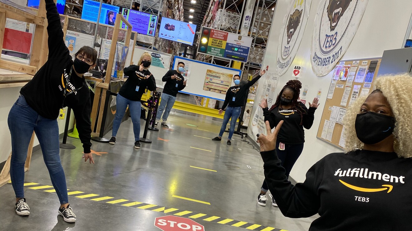 Six Amazon employees stand in an Amazon fulfillment center, preparing for Peak. All are wearing Fulfillment shirts, face masks, and are socially distanced.