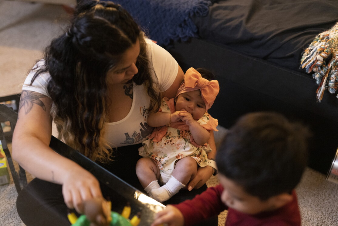 A woman holds a baby while a child plays nearby.