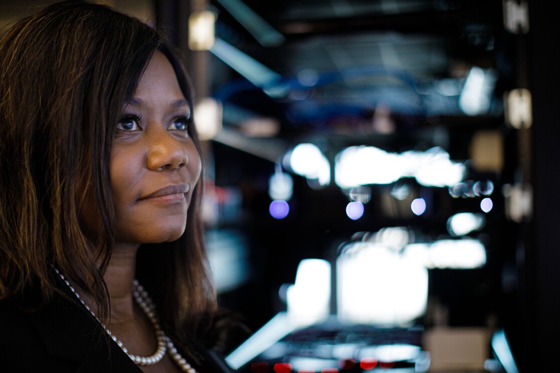 A woman in a black top and wearing pearls looks at the camera