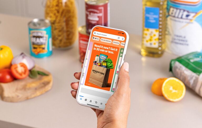 Amazon Now homepage appearing on a mobile phone with groceries in the background