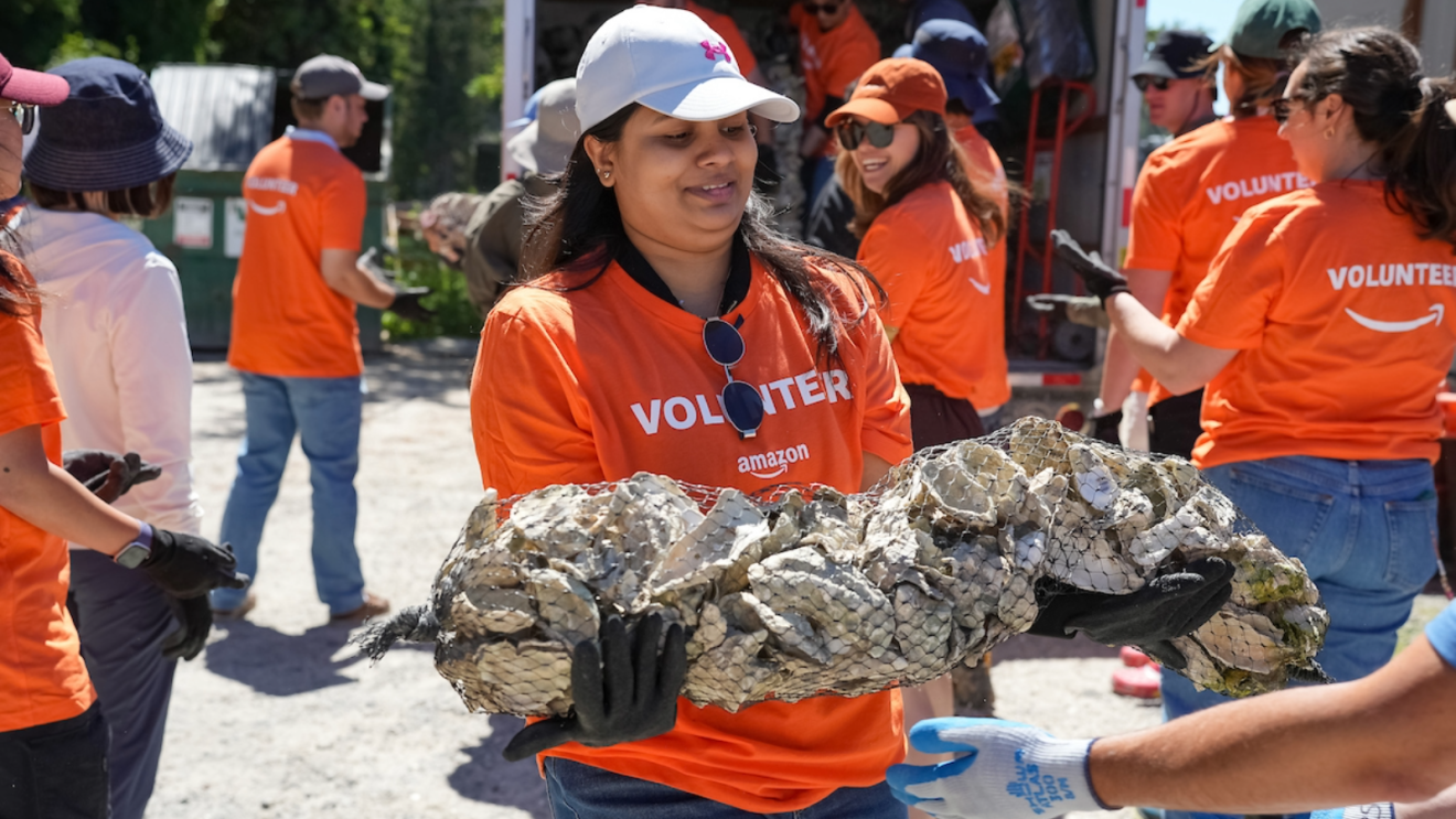 Amazon volunteers in orange shirts gather outdoors as a volunteer holds up oysters