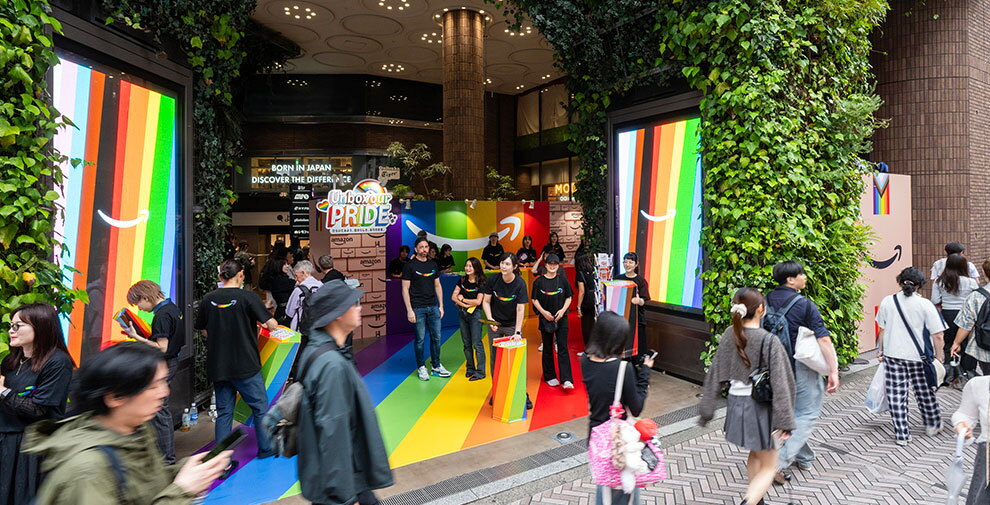 イベントが開催されいてる商業施設のエントランスと、その前の通りを歩く人々。イベントブースは床がレインボーカラーに塗られ、レインボーのサイネージも2画面設置されている。