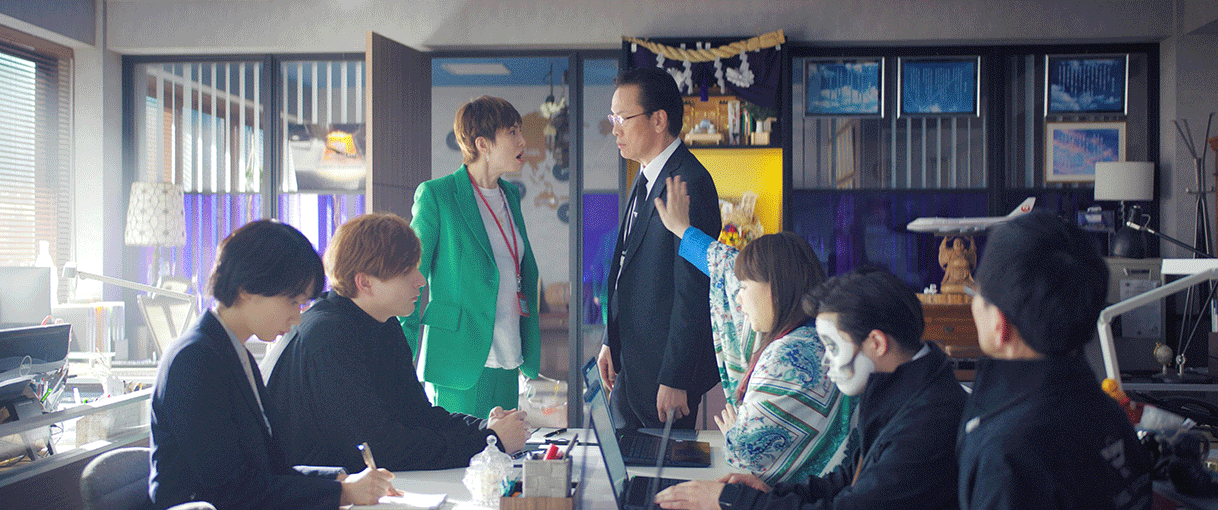 小さなオフィス中央のデスクに向かって作業する5人の人物。デスクの奥の2人の人物は立って対峙している。左側の派手な色のスーツの女性が右側の男性に話している。