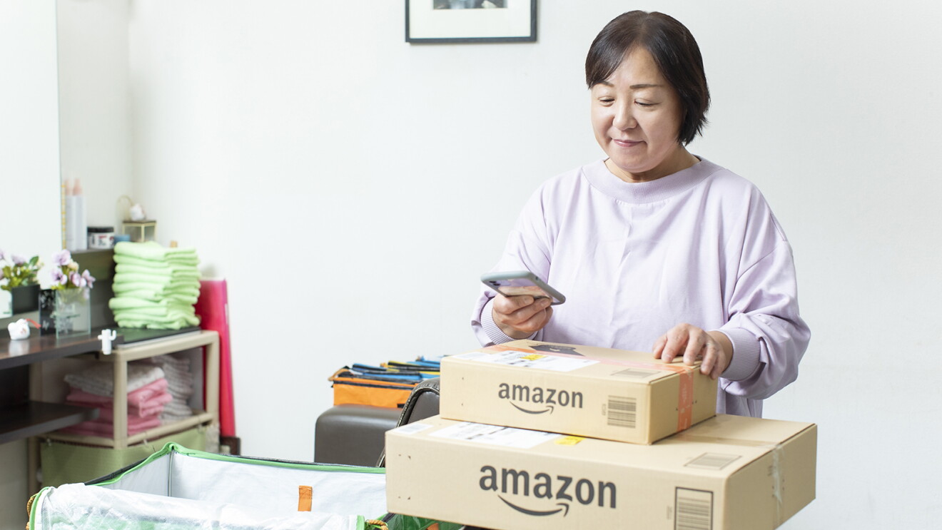 白い壁の室内。2つ重なったAmazonの段ボールを前に置き、スマートホンを操作する女性。部屋の隅の棚に、たくさんのタオルが畳まれておかれている。
