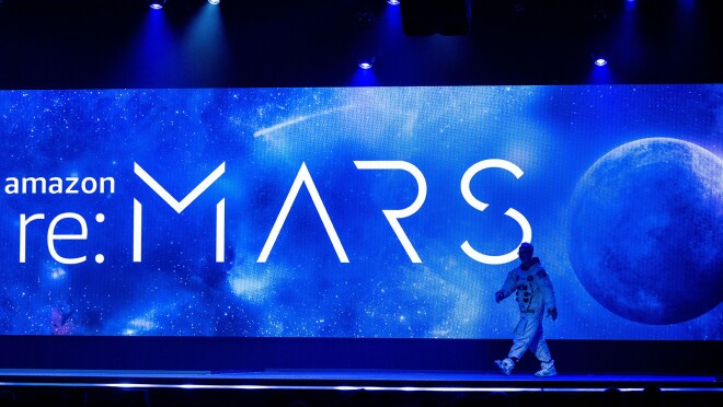 re:MARS 2022的舞台图像。一个穿着太空服的男人走上舞台。背景是一个大屏幕,上面写着“Amazon re:MARS”,上面有月球和太空的图像。