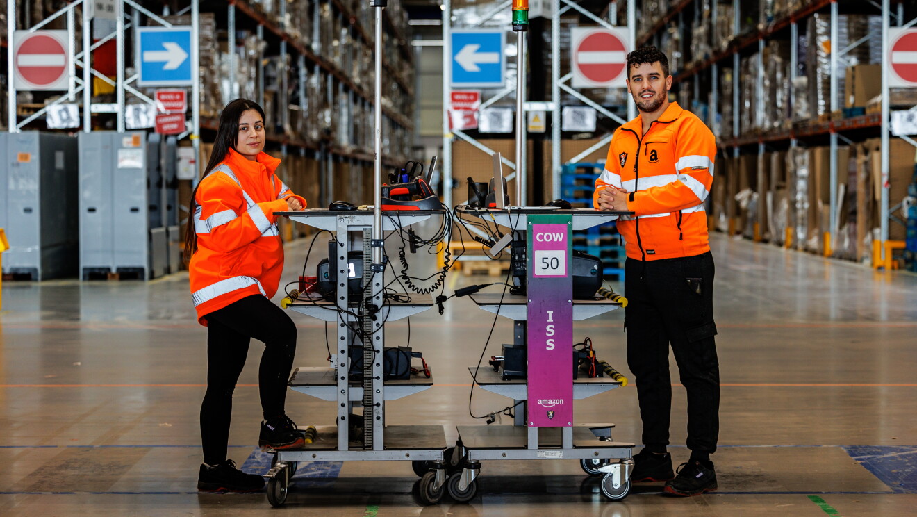 Miriam y Mario apoyados cada uno en una mesa alta transportable de Amazon. Los dos apoyan los brazo en la mesa. La mesa de él tiene un ordenador, la de ella tiene un ratón y un lector de código de barras. Los dos van con una chaqueta naranja que simula un chaleco de seguridad. De fondo el pasillo de un centro logístico. Miriam lleva el pelo largo por la cintura y es de color castaño. Él tiene el pelo corto pero la parte superior la lleva un poco más larga y la tiene rizada. Tiene los ojos claro, barba de dos días y un piercing en el labio inferior. De fondo las estanterías de un centro logístico.
