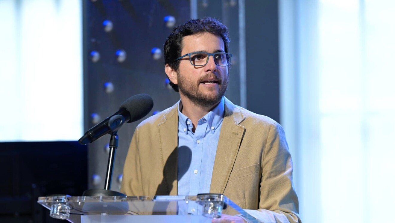 Andrés Rodríguez, responsable de Servicios a Vendedores, Amazon Marketplace para España, aparece en un atril transparente con un micro negro. Está de pie, con las manos en el atril. Lleva pelo corto y barba de dos días, camisa azul y americana marrón. 