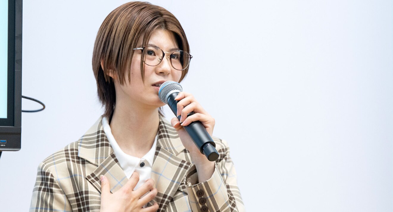 白い背景を背に、眼鏡をかけた人物が左手にマイクを持って話している。ベージュ地に茶色とライトブルーのチェック柄のジャケットと白いシャツを着用、茶髪のショートカット。
