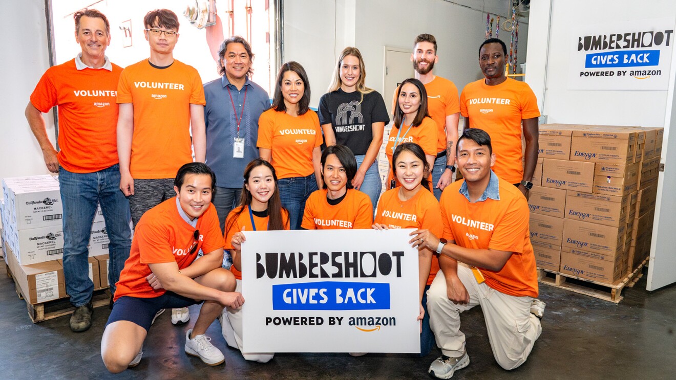 Amazon and Bumbershoot volunteers holding a 'Bumbershoot Gives Back' sign in front of delivery boxes.