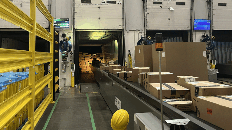 An image of boxes on a conveyer belt in an Amazon fulfillment center leading up to an open truck where there are a lot of boxes stacked.