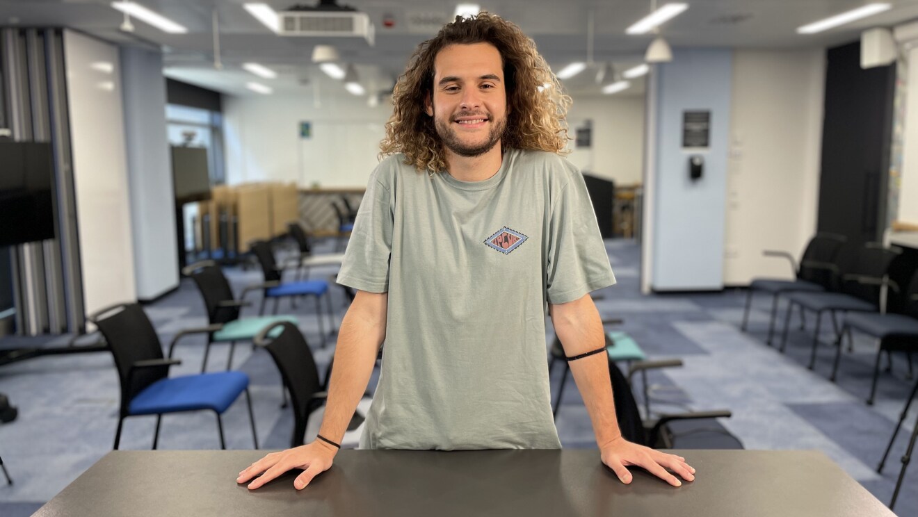 Andrés de Cossío mirando a cámara con los brazos apoyados en una mesa de color gris. Andrés tiene el pelo largo rizado por el hombro y suelto. Camiseta de algodón de manga corta y de color gris. En cada muñeca lleva una goma de pelo. Tiene también barba y perilla de dos días. De fondo se ve una sala con sillas y mesas de tonos azules y verdes y una pizarra blanca de fondo.