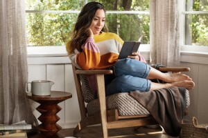 A woman lounges in a chair, with her feet tucked to the side, reading a new Amazon Kindle Paperwhite. 