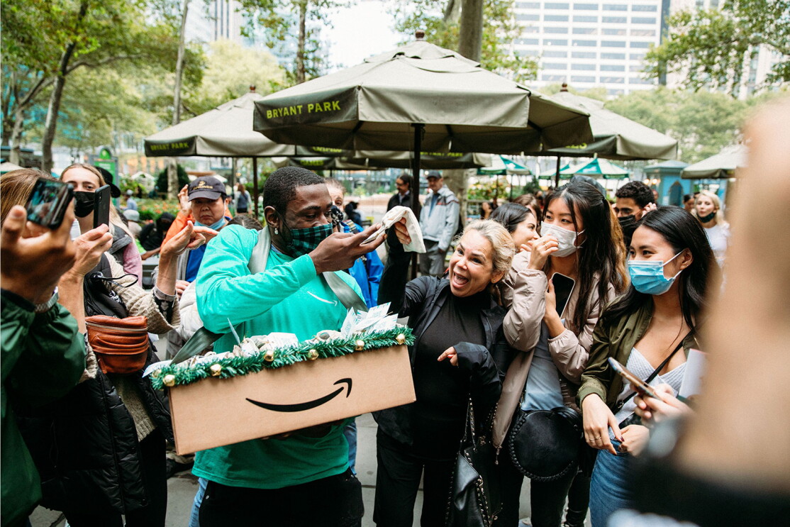 An image of a man passing out treats to an excited crowd. The box with the treats has the Amazon logo on it. The man is wearing a green Amazon shirt and a black and green checkered mask. 