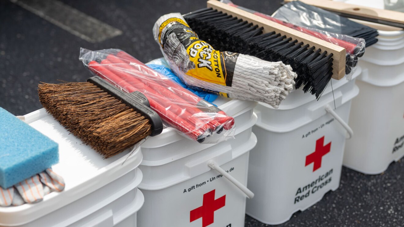 An image of several white pales lined up. The pales have the Red Cross logo on them, and all of them have various cleanup items like brooms, sponges, and gloves on them.