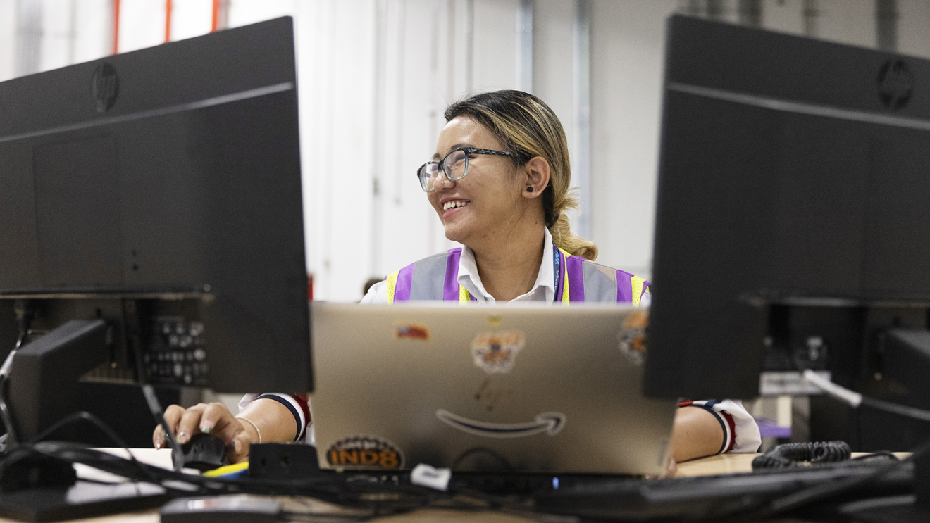 A photo of an Amazon employee sitting at a desk and working on a laptop device that is connected to two large monitors.