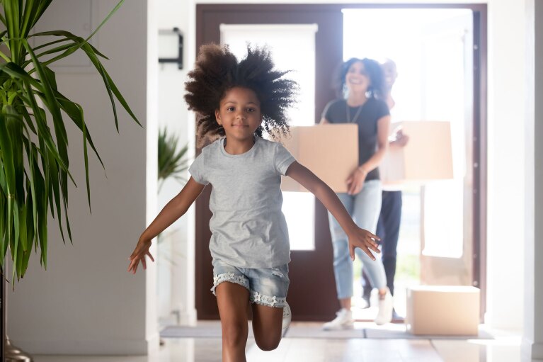 A little girl runs into a house.  Adults carrying moving boxes into the home are out of focus behind her.