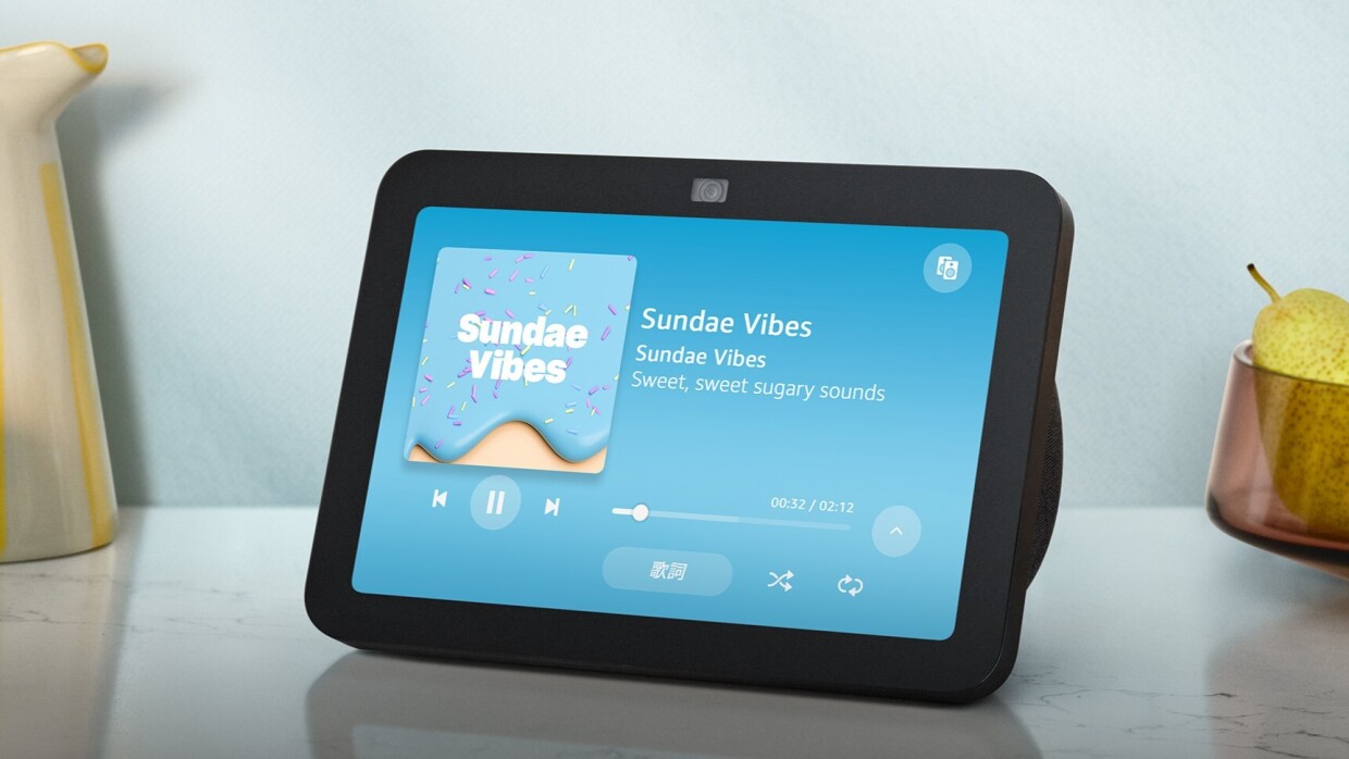 果物と水差の間に黒いフレームのEcho Show 8がテーブルの上に置かれている。画面には青い背景に音楽のジャケットと一時停止マークなどが表示されている