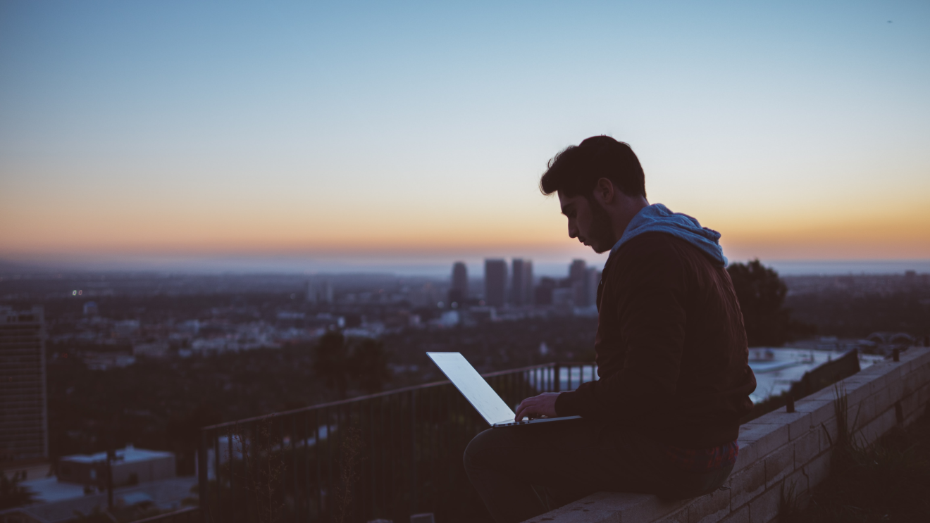 Man sat on a wall typing on his laptop 