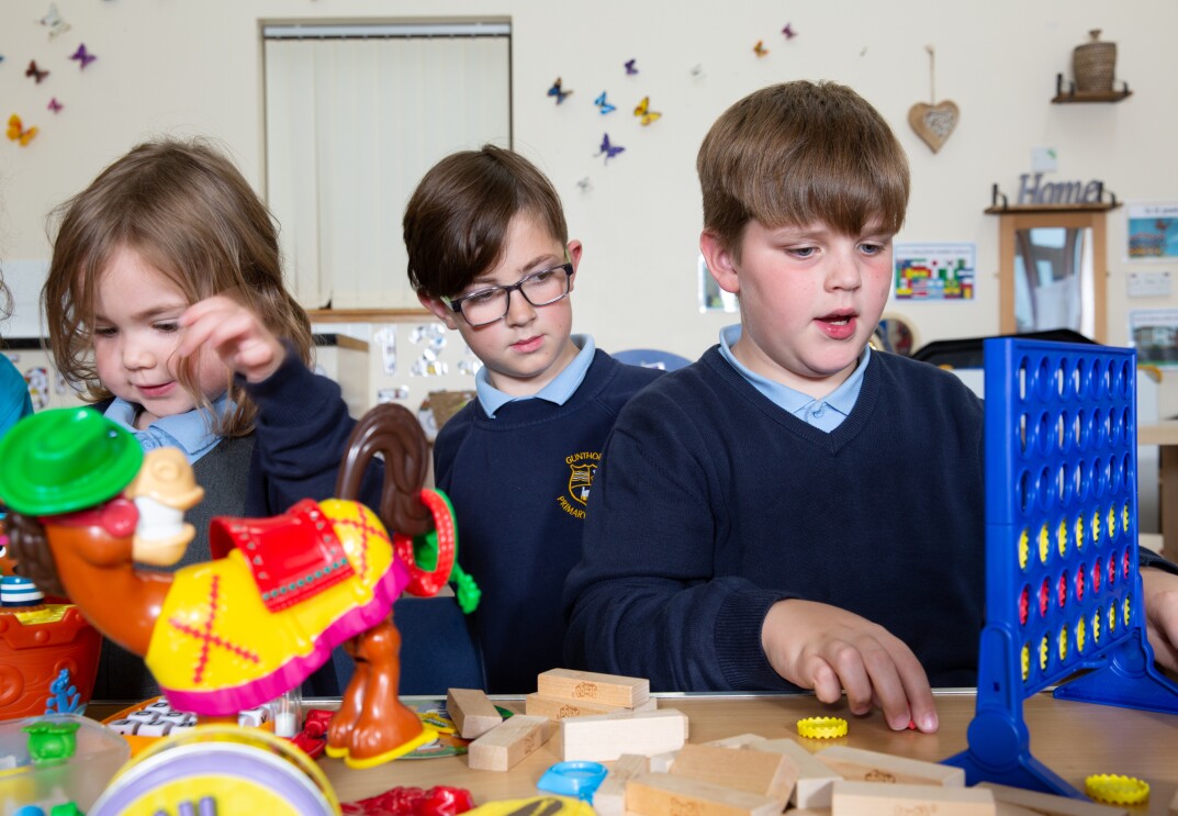 Three school children playing various games on a table, there is Connect Four, Buckaroo, Jenga and others.