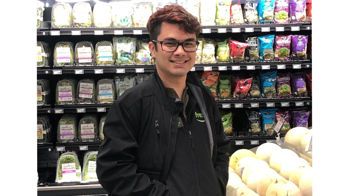 Eli smiles for the camera in front of the packaged salads and produce section of the grocery store. He wears a black Amazon Fresh zip-up and black-framed eyeglasses.