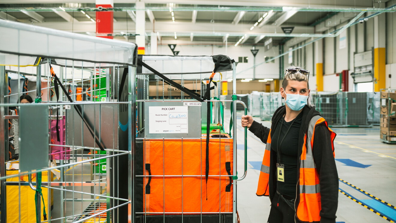 Elisabetta indossa una mascherina chirurgica sul volto. Si trova all'interno di un magazzino Amazon e con la mano destra tra trasportando un carrello con dei pacchi Amazon. 