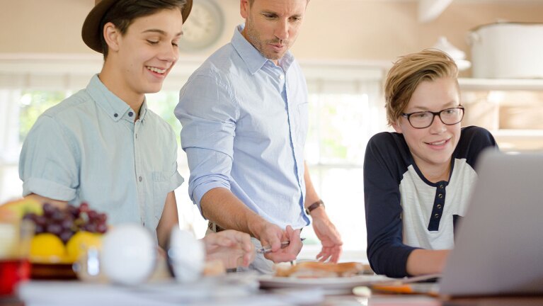 A father and two sons gather around a laptop on the kitchen table.