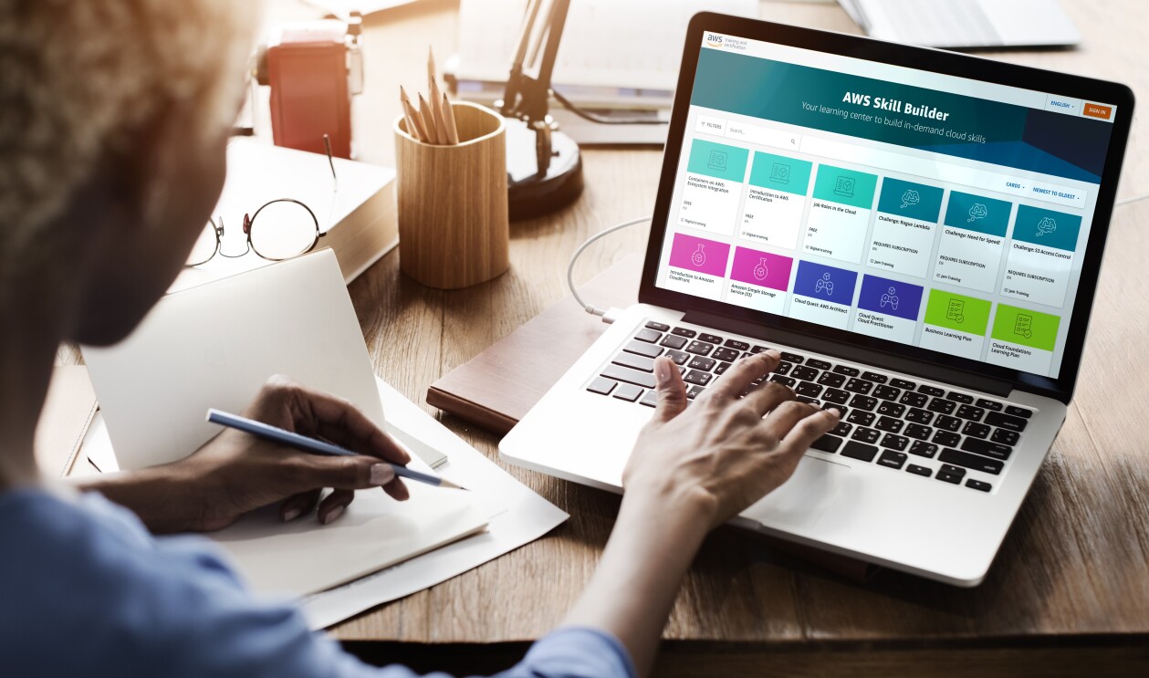 La pagina AWS Skill Builder aperta su un laptop. Una donna di spalle sta digitando sul computer con la mano destra, mentre con la sinistra prende appunti su un'agenda