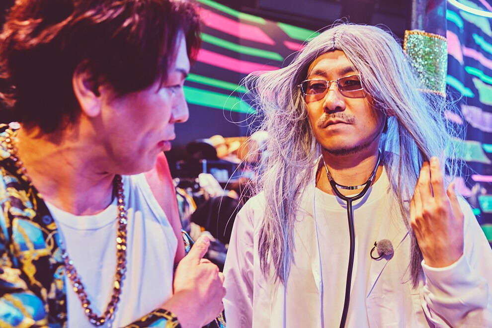 黒地に黄緑とピンクの横縞が入ったセットで向かい合う2人の男性。左側の男性は派手なシャツと金のチェーンネックレス、右側の男性は長い白髪のかつらと白衣、聴診器をつけている。