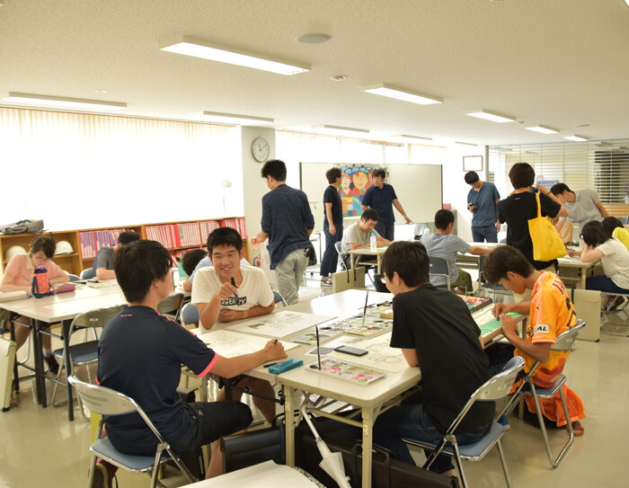 広い室内に集まる人々。デスクを集めて複数のグループを作り、絵を描いている。