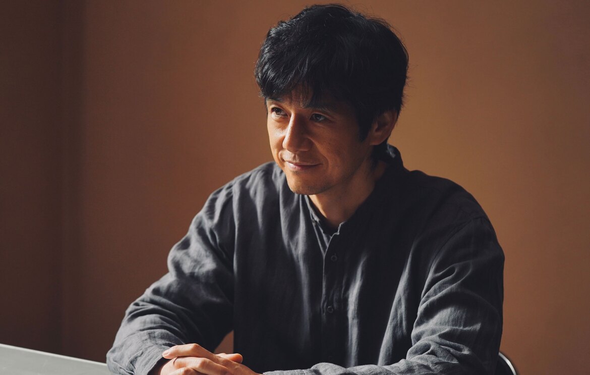 茶色い壁を背景に、グレーの柔らかそうな素材のシャツを着た人物の上半身。机のうえで両手を組み、笑みを浮かべている。