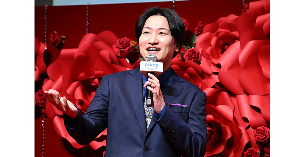 赤いバラの花の大きなオブジェと生花の赤いバラが飾られた赤い壁を背に、マイクを持って話すスーツ姿の男性。右手を前に差し出している。