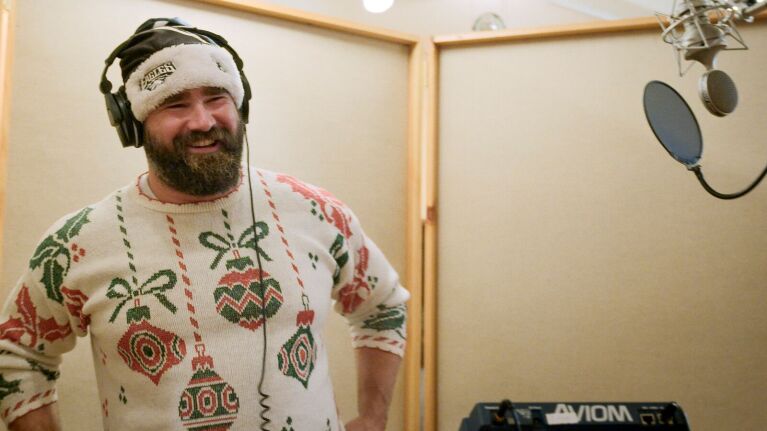 NFL center Jason Kelce laughing in a recording studio wearing an Eagles beanie and a Christmas sweater.