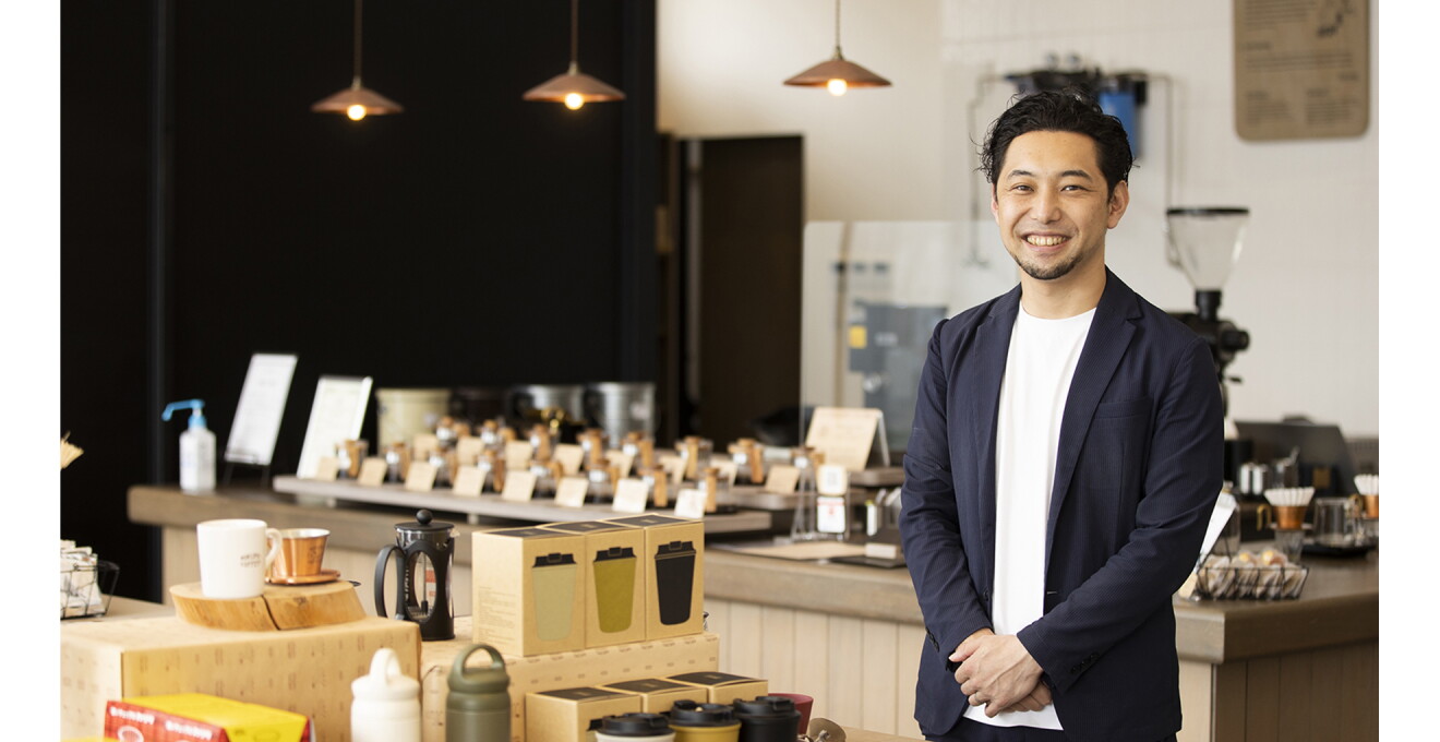 多くのコーヒー豆やコーヒーグッズの並ぶ店舗を背景に井田さんの写真
