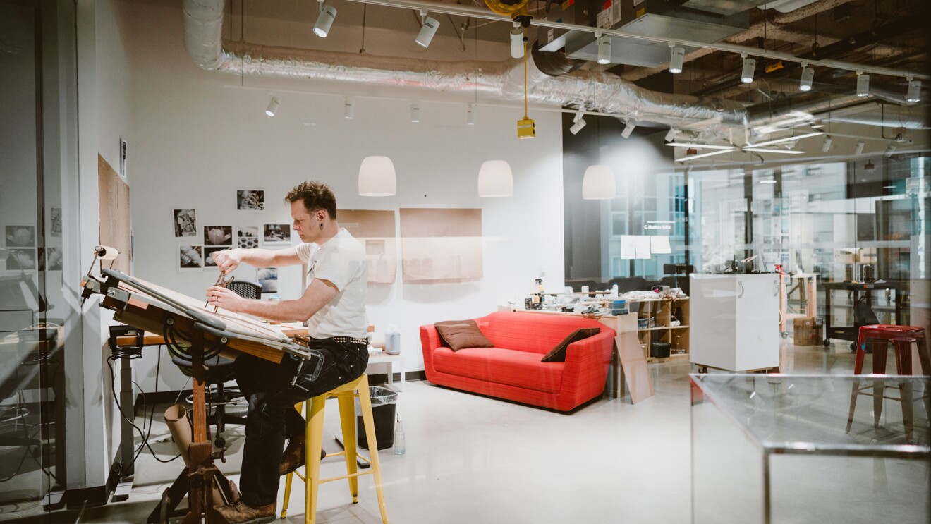 An artist sits at a raised desk, working on a project within an Amazon office building