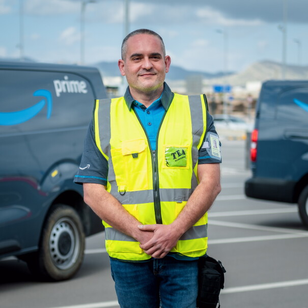 Juan Antonio Carrasco, reconocido como Driver Hero. Está posando mirando a cámara, con las manos cruzadas delane y de fondo furgonetas de color gris con el logo, sonrisa de Amazon, en azul y la palabra Prime en blanco. Él está con las manos cruzadas delante, un polo azul de manga corta y una chaleco de color amarillo. Tiene el pelo corto negro y blanco. 
