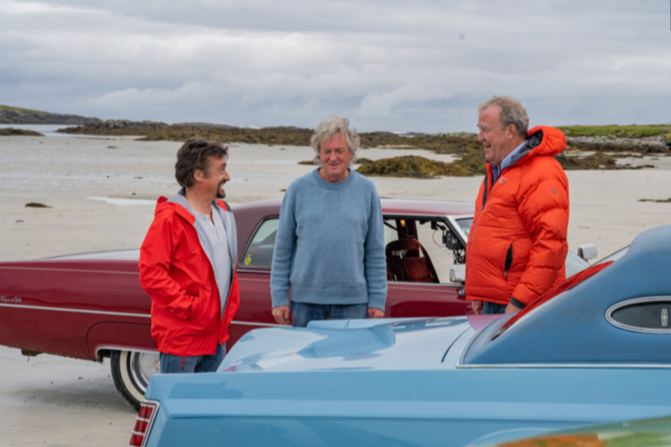 Three men (Richard Hammond, James May and Jeremy Clarkson) stand next to two cars on a beach