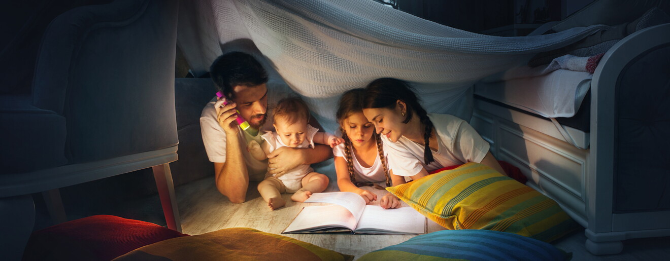A family sits in a homemade fort, reading a book together by flashlight.
