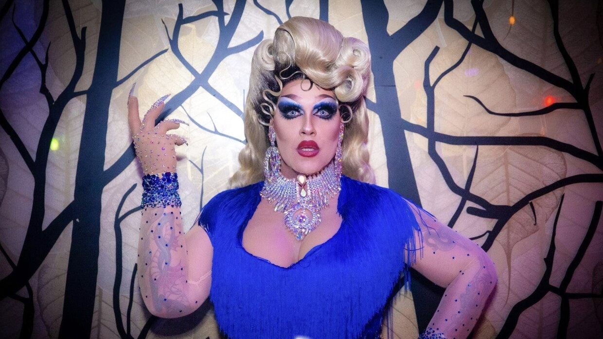 Primo piano di Lucrezia Borkia, drag queen dipendente di Amazon. Indossa un vestito blu elettrico con le maniche in tulle e delle frange, una parrucca bionda e una collana di diamanti. Sullo sfondo, una parete con degli alberi disegnati.