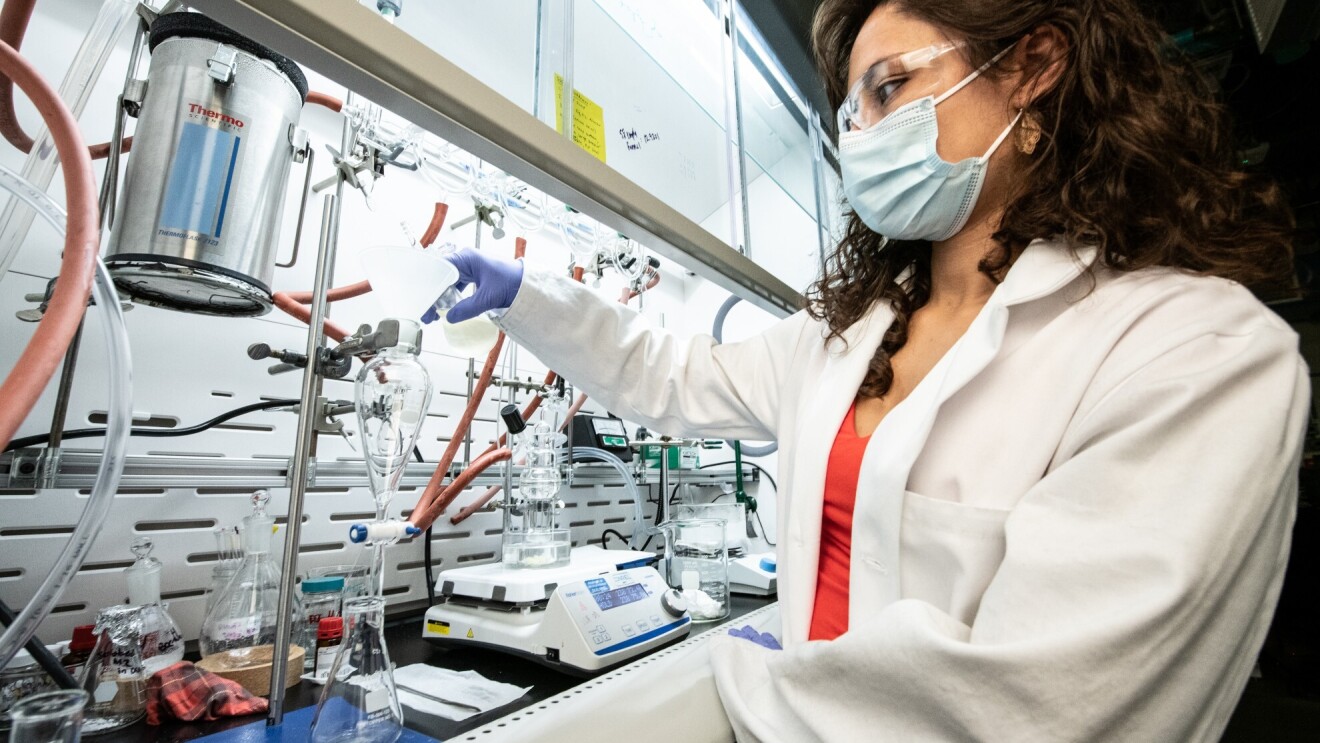 A woman scientist wears a mask and pours liquid into a glass beaker.