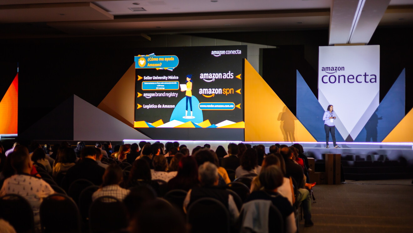 Mujer realizando una ponencia delante de un cartel que pone Amazon Conecta. De fondo también se ve una proyección con información con infografías respondiendo a la pregunta: ¿cómo se ayuda Amazon?