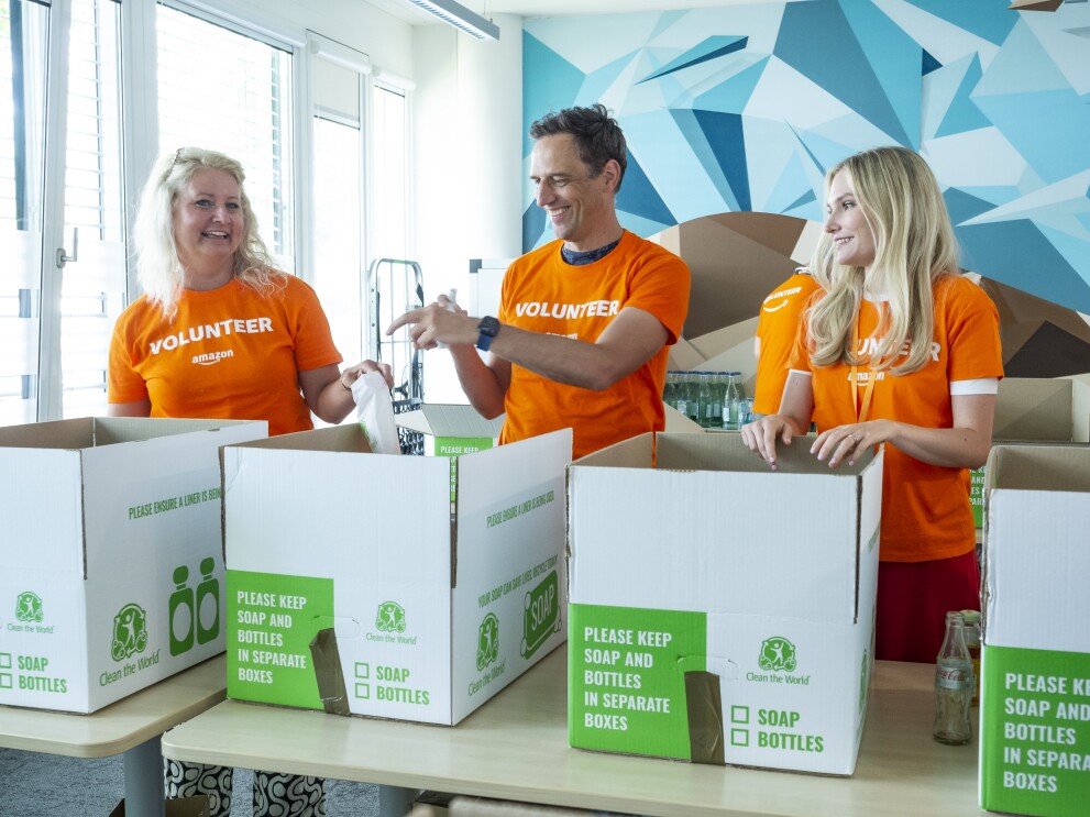 Rocco Bräuniger mit zwei Amazon Mitarbeiterinnen in orangenen Volunteer T-Shirts beim Packen von Hygienesets für bedürftige Geflüchtete.