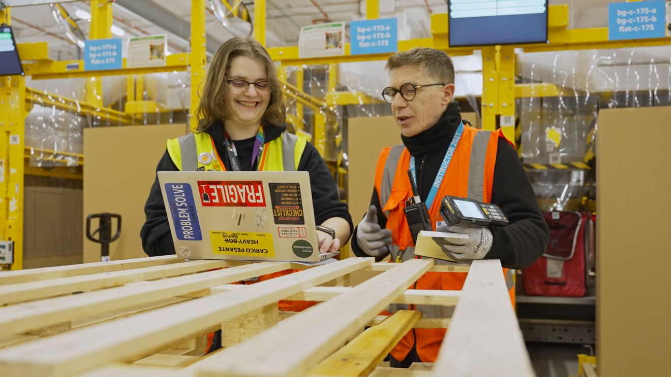 A young woman and a man with yellow and orange vests are looking at something on a laptop screen inside a fulfillment center