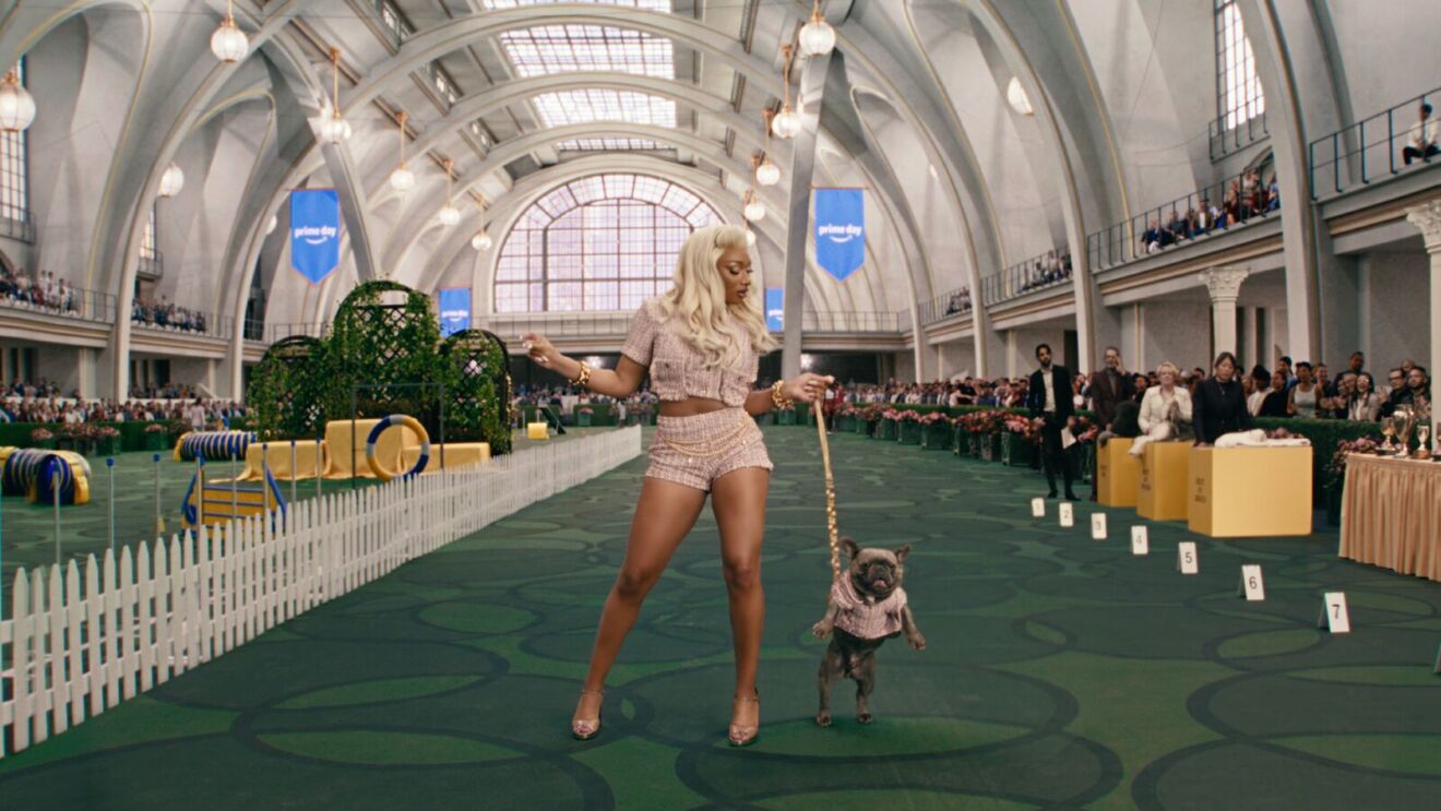 An image of Megan Thee Stallion walking her dog at a dog show. She is walking along the turf grass wearing a tweed set with shorts and a cropped top.