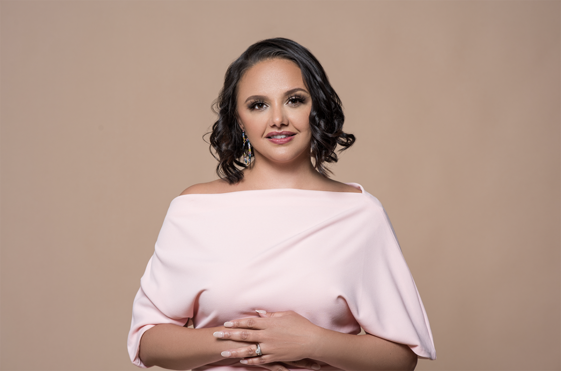 An image of a woman smiling for a photo in front of a light brown background. 