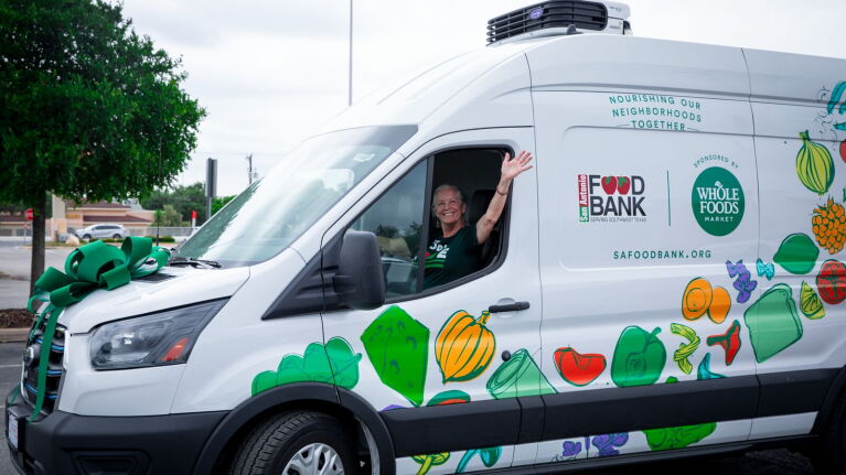 Food bank driver in green shirt waves from white refrigerated truck with 'Nourishing Our Neighborhoods Together' slogan and produce graphics