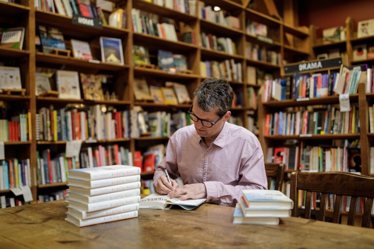 An author sits at a table and signs his book in a bookstore, surrounded by shelves of other book titles.