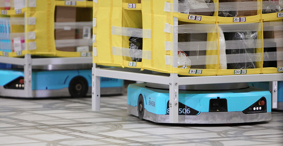 グレーの床に、平たい水色の自動搬送ロボットがあり、その上に黄色い収納棚がある。それがいくつも並んでいる