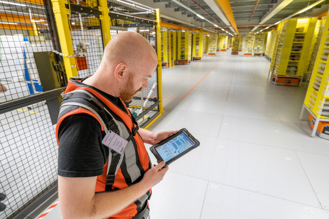 Ein Mitarbeiter mit Sicherheitsweste und Tablet steht in einem Bereich mit Transportrobotern.