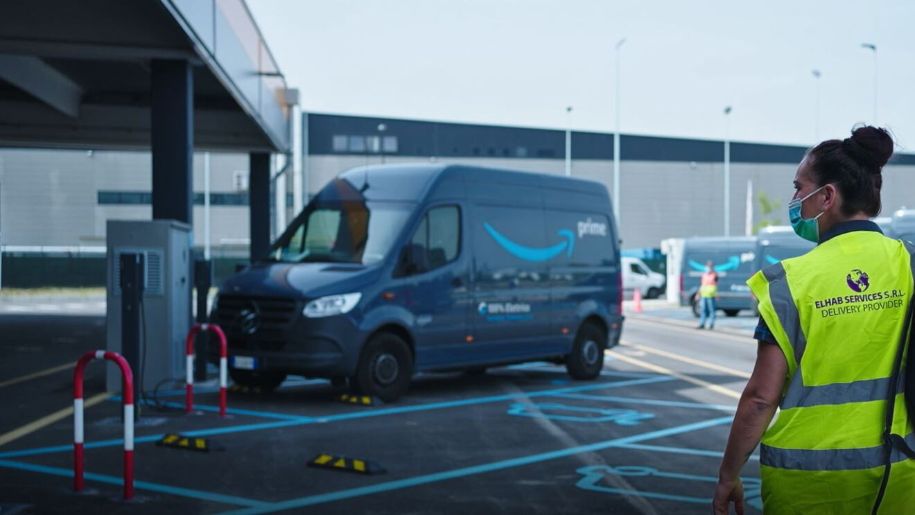 Una donna girata di spalle con un giubbino di sicurezza giallo a strisce catarinfrangenti e la scritta Elhab Services. Sta camminando in un parcheggio in cui si vedono dei furgoni con il brand Amazon Prime. La donna ha una coda alta e porta uno zaino sulla spalla destra. 