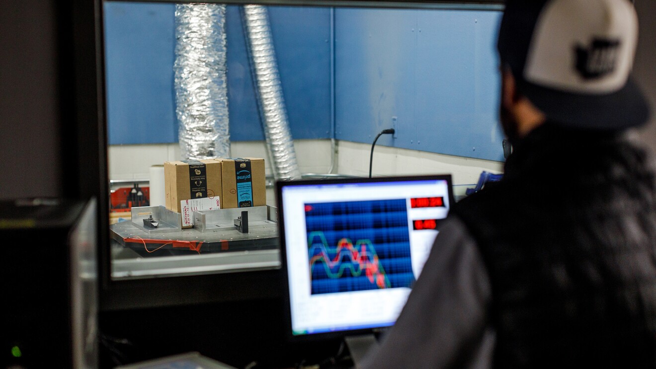 Aman sits behind a computer in a packaging lab and watches while tests are executed